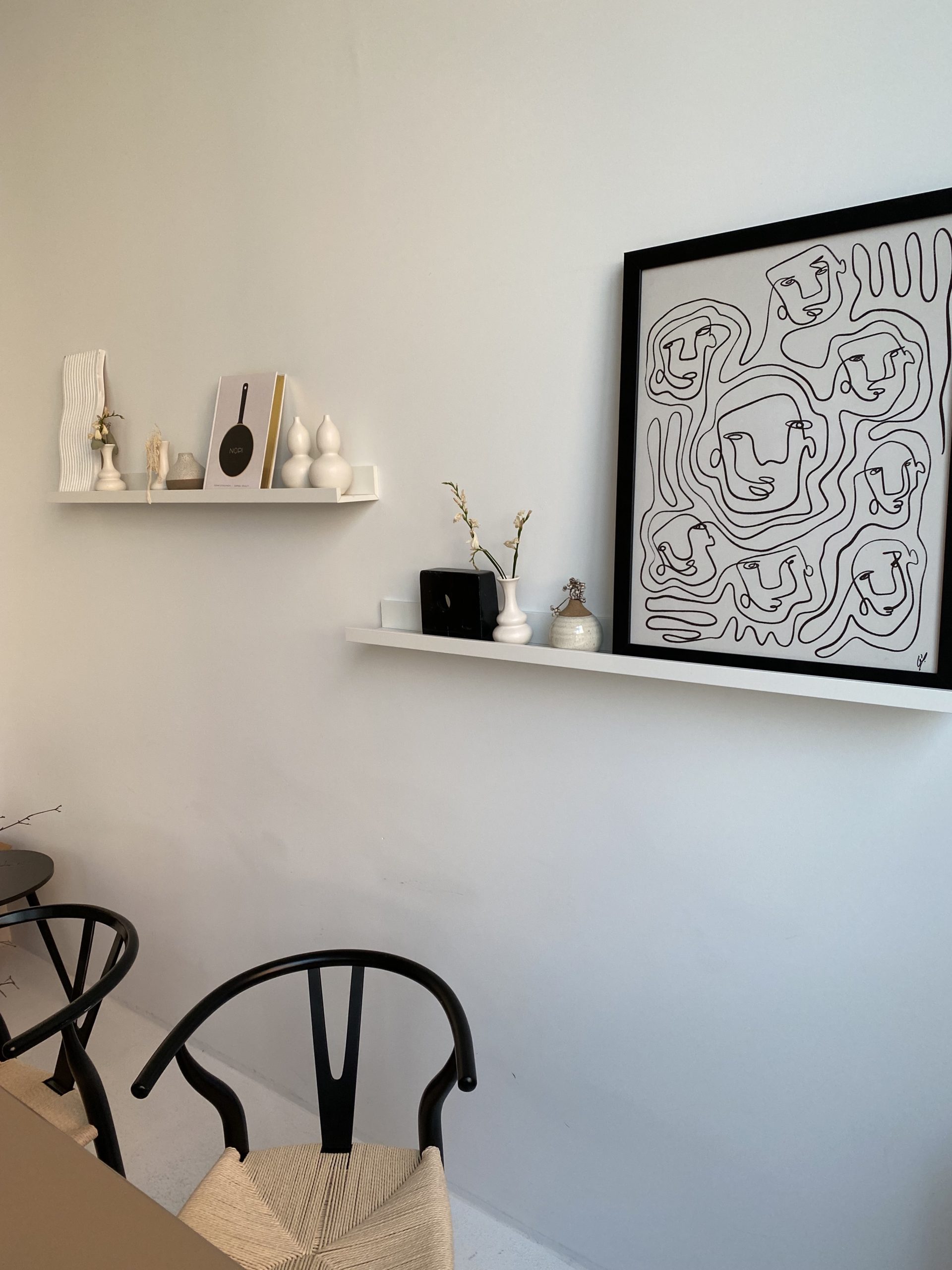 Set against a blank white wall. Tjere is a black and white photo on a shelf next to white trinkets (a mini vase, a small book). There are two wicker chairs with black frames.