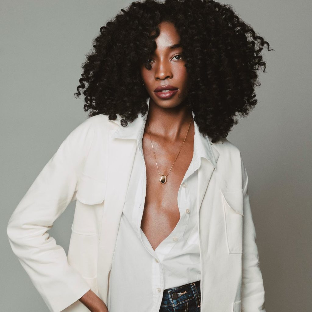 Black woman with thick curly hair. She is wearing a white button up shirt and a white blazer. Striking a bold pose