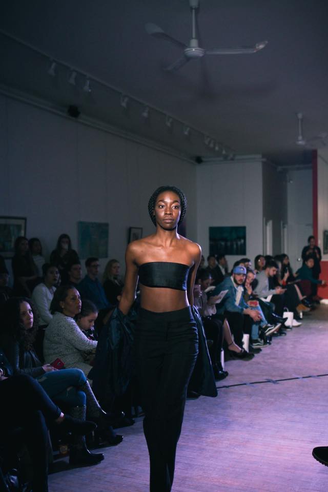 Black woman walking a runway. She is wearing a matching black crop top with black pants. There is an audience to her left