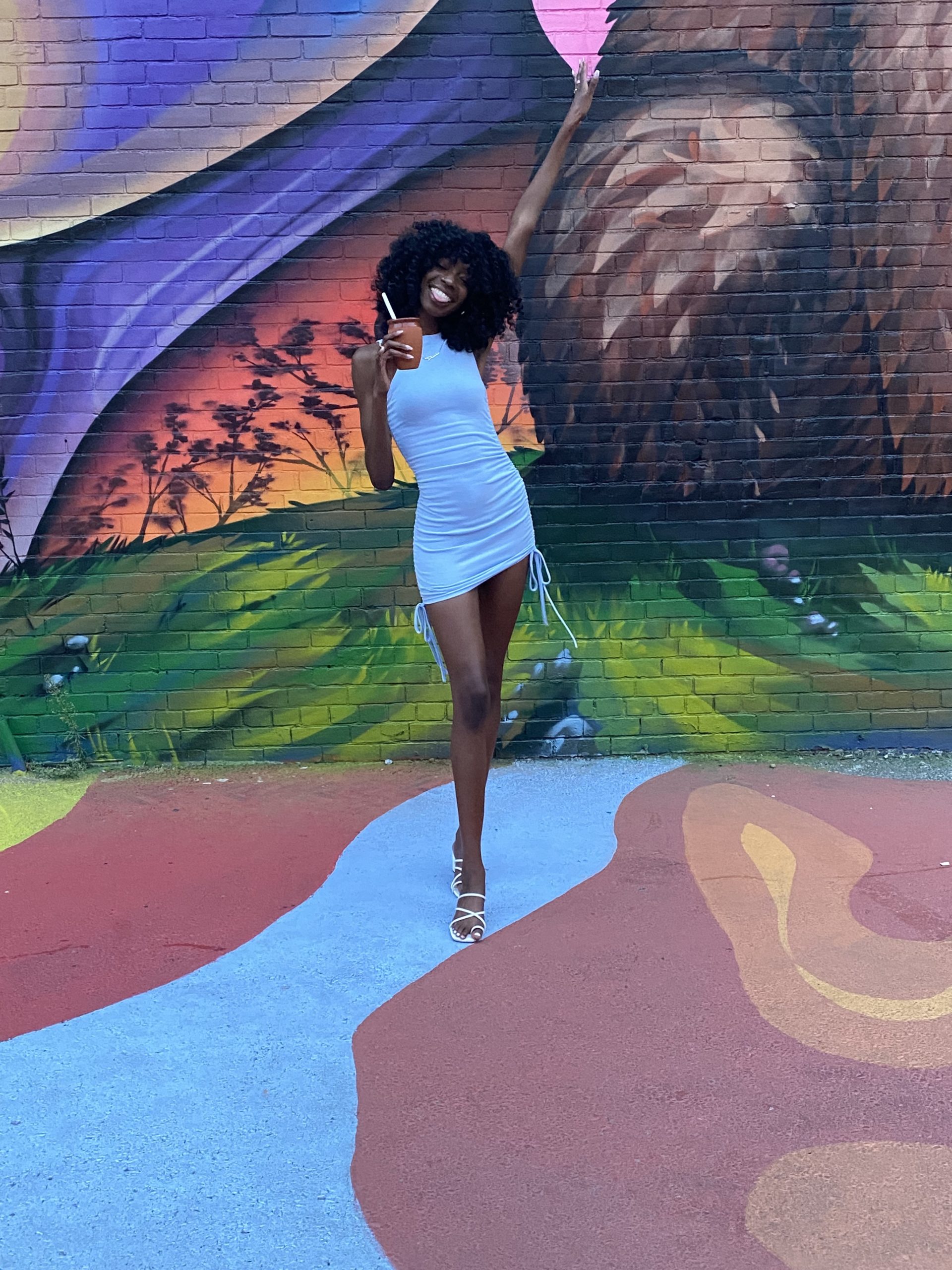 Tall black woman wearing a bright blue dress with big curly hair. She is smiling and standing in front of a colourful grafitti wall and holding a brown cup