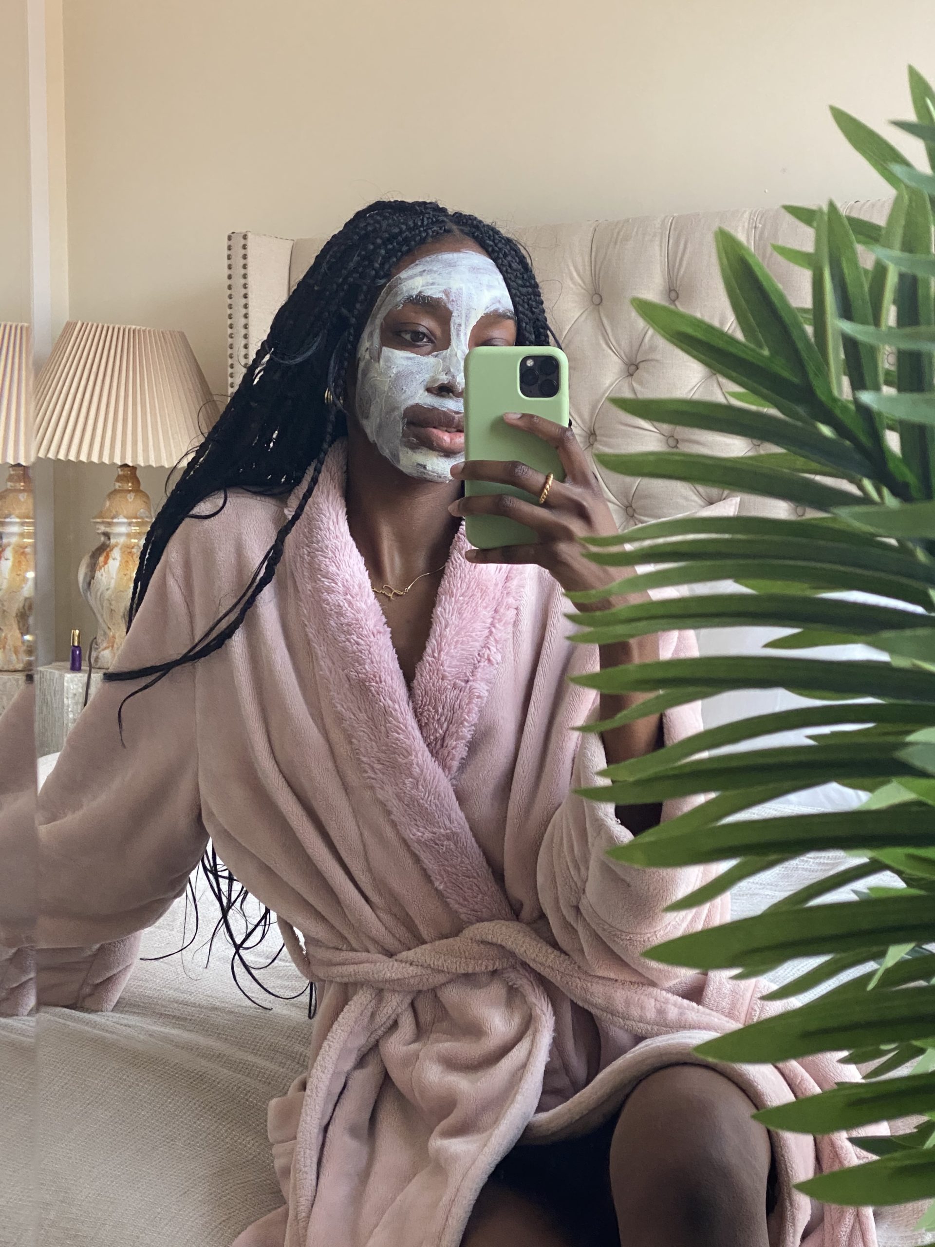 Black woman with white face mask and long braids. She is sitting on a bed in a fluffy pink robe taking a selfie of herself. There is a green plant in the right corner